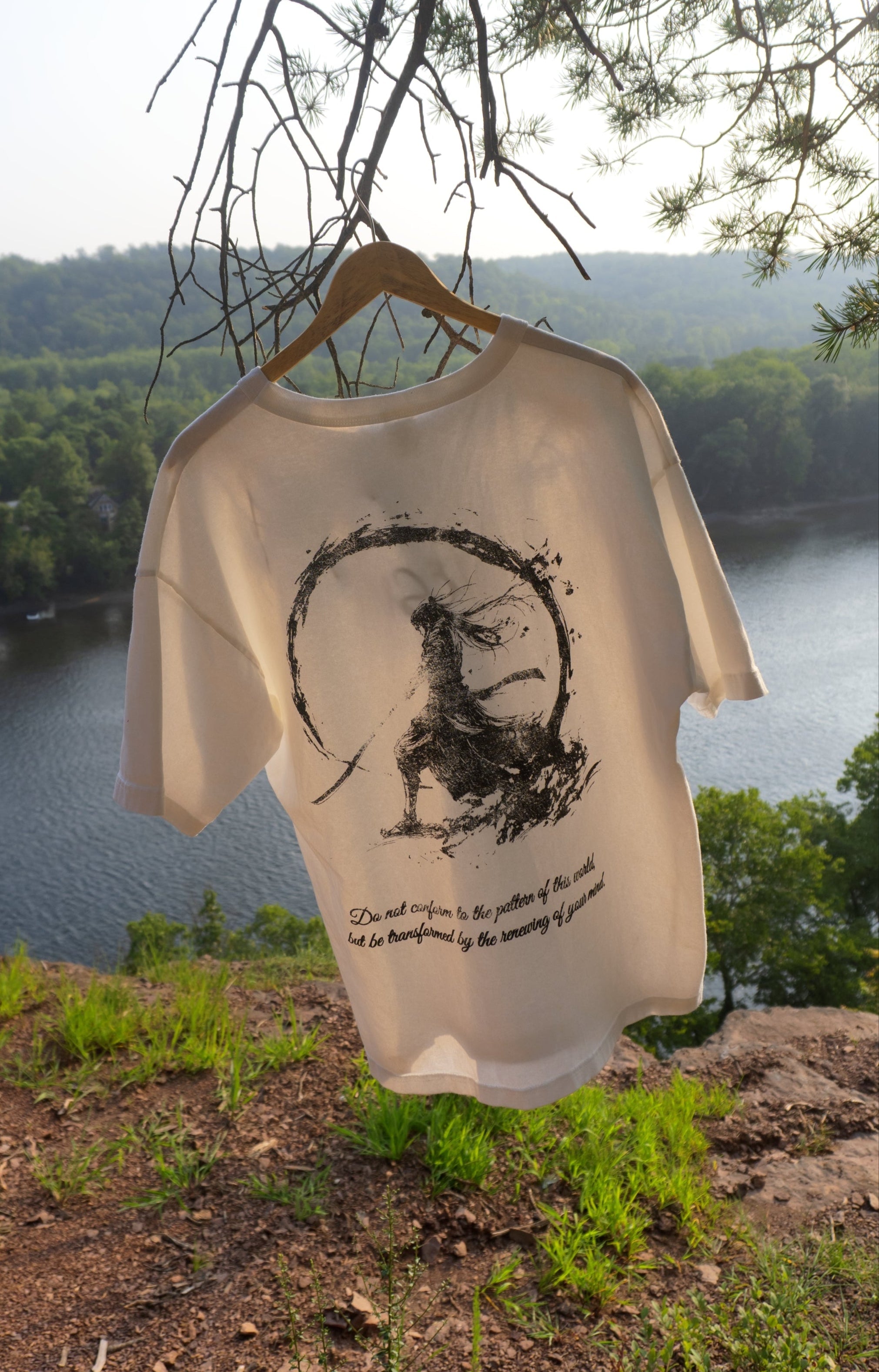 White t-shirt with black graphic design hanging on a tree branch over a scenic view of a lake and forest.